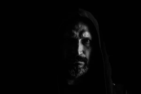 Brutal man with a gray beard in the hood with sharp shadows on a black background. Copy space. — Stock Photo, Image
