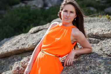 Turuncu uzun elbiseli genç kadın açık havada. Kayalar ve çim arka plan.