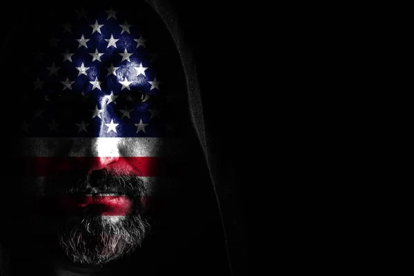 Жестокий человек с седой бородой, с флагом США на лице, в капюшоне с острыми тенями на черном фоне. Копирование пространства — стоковое фото