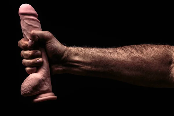 Adult present. Artificial penis in mens hand on dark background.