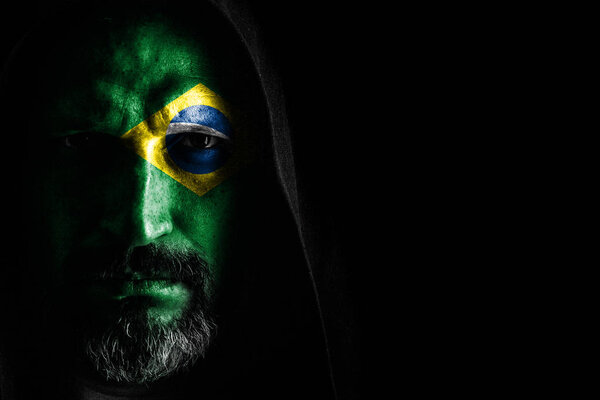 Жестокий человек с седой бородой, с флагом Бразилии на лице, в капюшоне с острыми тенями на черном фоне. Копирование пространства
