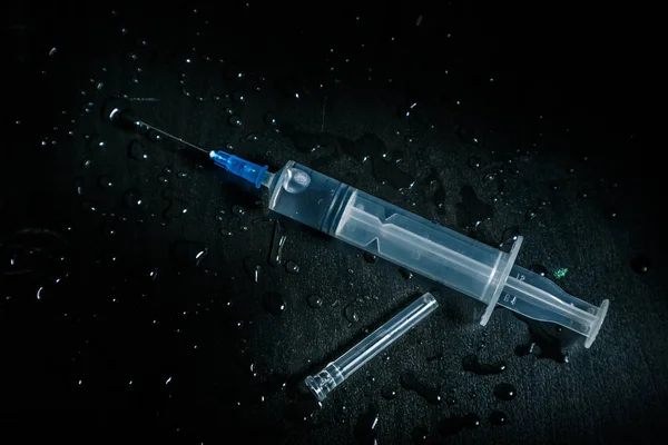 A syringe with a clear liquid lies on a dirty dark background covered with drops. — Stock Photo, Image