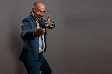 Sakallı, beyaz gömlekli ve mavi kravatlı bir ceketgiyen kel orta yaşlı bir adam, kirli gri bir arka plan üzerinde onay işareti olarak başparmak ları yukarı yaslar.