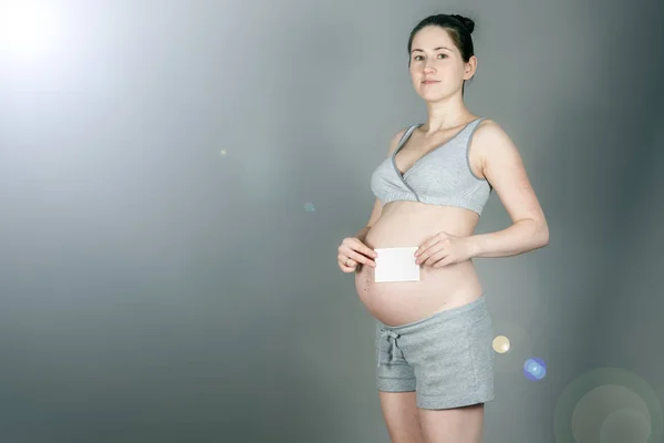 Une jeune femme enceinte en costume gris modeste tient une feuille de papier blanche qui ressemble à une échographie sur un fond gris sale . — Photo