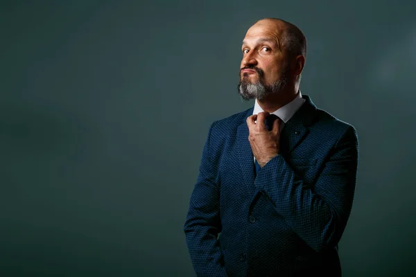 Bald middle-aged man with a beard, in a jacket with a white shirt straightens his blue tie on a dirty gray background