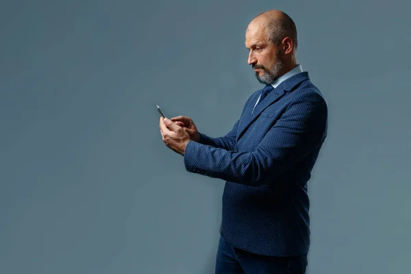Bald middle-aged man with a beard, in a jacket with a white shirt and blue tie holding a mobile phone on a dirty gray background