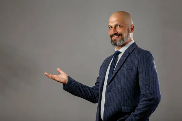 Hangi konu yerleştirmek için açık bir avuç içi ile klasik bir takım elbise sevimli genç kel sakallı işadamı. Kopyalama alanı ile gri arka plan