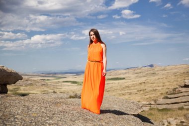 Turuncu uzun elbiseli genç kadın açık havada. Kayalar ve cennet arka plan.