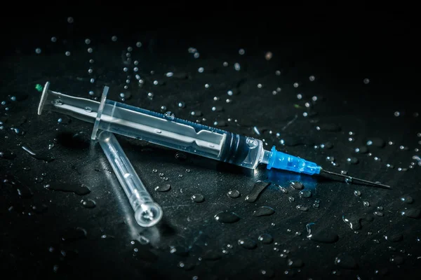 Açık sıvı olan bir şırınga, damlalar ile kaplı kirli karanlık bir arka planda yatıyor.