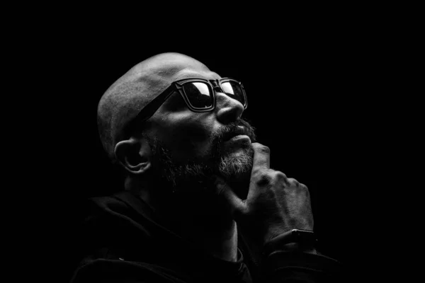 Pensive bald man with a gray beard in sunglasses on a dark background Stock Picture