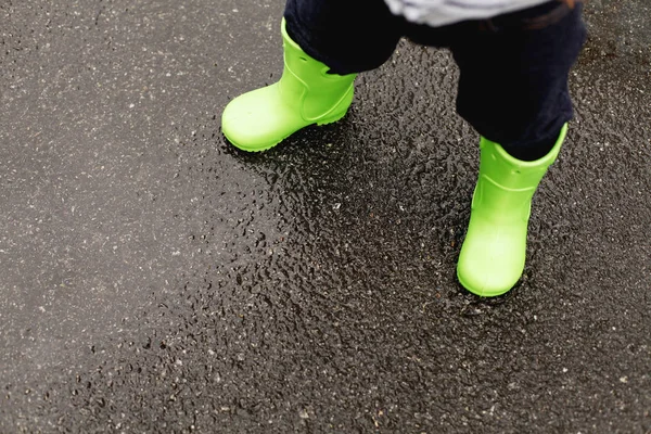 Closeup of child legs in green gum boots standing on wet asphalt