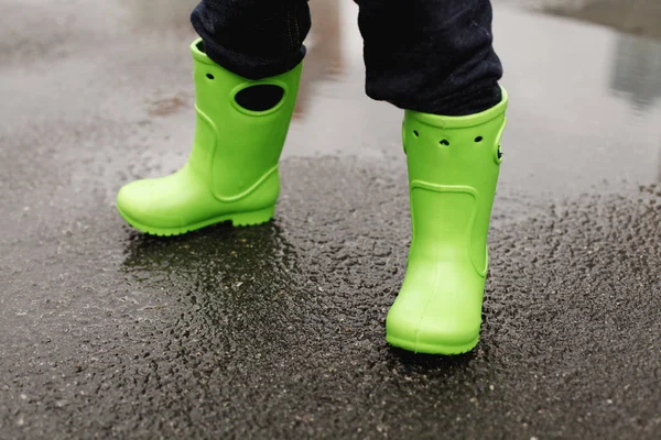 Closeup of child legs in green gum boots standing on wet asphalt