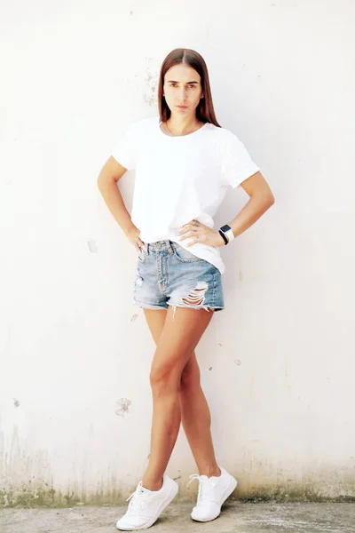 Fitness young girl with long legs in denim shorts and sneakers posing against the white wall, simple attire