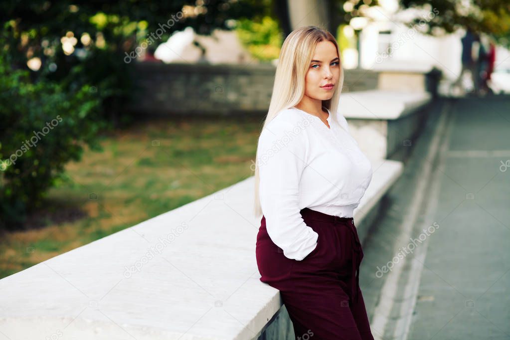 Young stylish woman wearing white blouse and burgundy color pants on the city street in autumn. Casual fashion, elegant look. Plus size model. Fall fashion