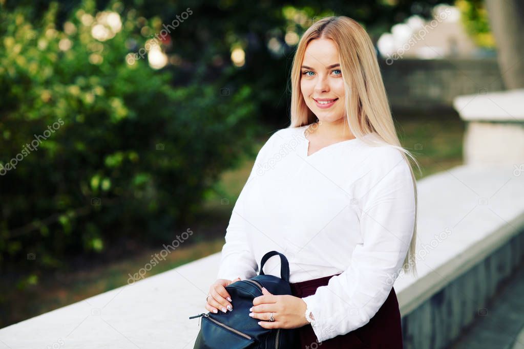 Young stylish woman wearing white blouse and burgundy color pants on the city street in autumn. Casual fashion, elegant look. Plus size model. Fall fashion