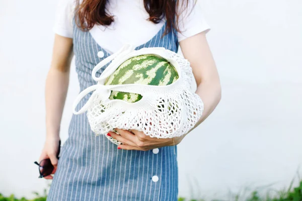 Young woman in hat, denim dress posing with watermelon in knitted bag