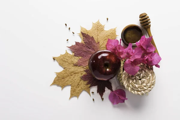 Rosh Hashanah Jewish holiday concept - red apple, wood dipper, saucer of honey on autumn maple leaves, white background with pink flowers, wicker baskets and cinnamon. Traditional holiday symbols
