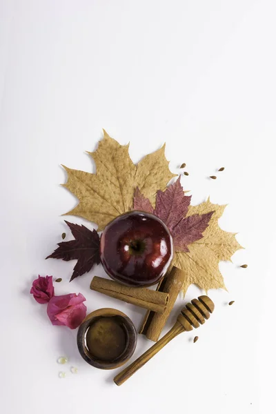 Rosh Hashanah Jewish holiday concept - red apple, wooden dipper, saucer of honey on autumn maple leaves, white background with pink flowers and cinnamon. Traditional holiday symbols