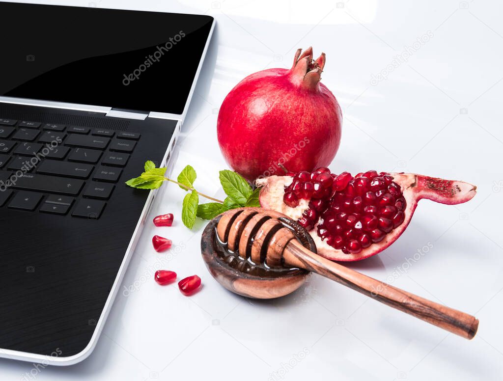 Rosh Hashanah Jewish holiday concept - red pomegranate and slice, honey in wooden saucer with honey dipper, green leaves and part of laptop. Traditional holiday symbols