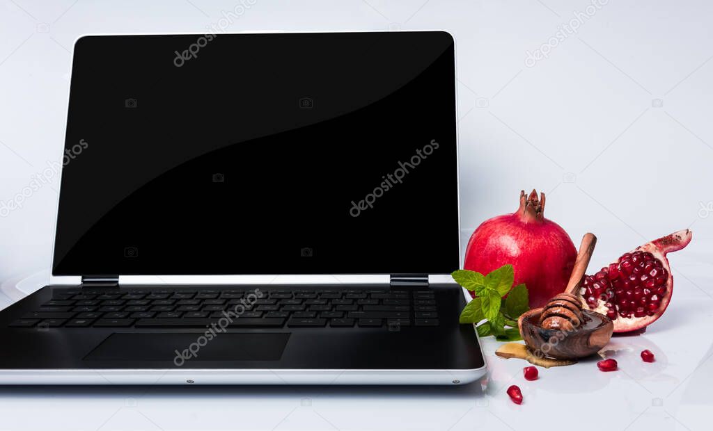 Rosh Hashanah Jewish holiday concept - red pomegranate and slice, honey in wooden saucer with honey dipper, green leaves and laptop. Traditional holiday symbols