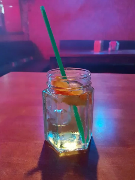 A glass of cocktail with straw, lemon and cubes of ice placed on table in the night bar, restaurant while disco lights shining on cocktail.