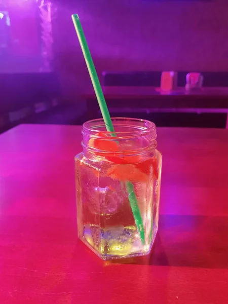 A glass of cocktail with straw, lemon and cubes of ice placed on table in the night bar, restaurant while disco lights shining on cocktail.