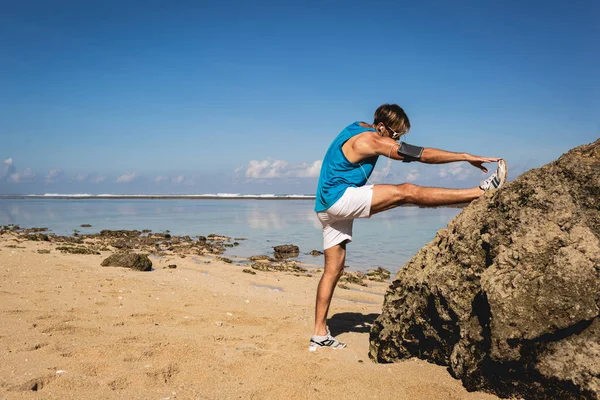 运动男子伸展腿近岩石在海滩上 — 图库照片