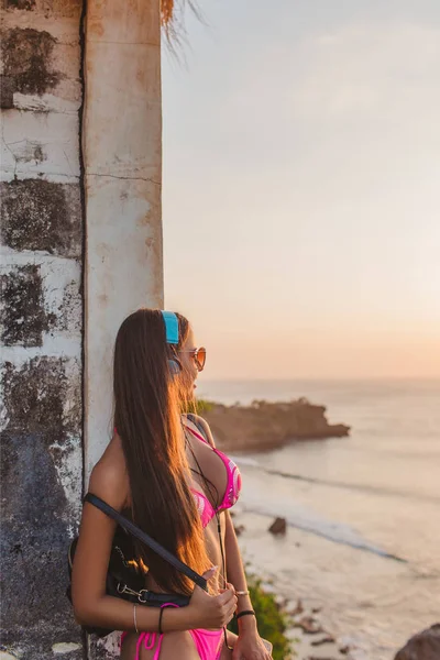 ヘッドフォンと壁にもたれして海でピンクのビキニで魅力的な女の子の側面図  — 無料ストックフォト