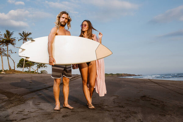 couple standing with surfboard on beach in bali, indonesia