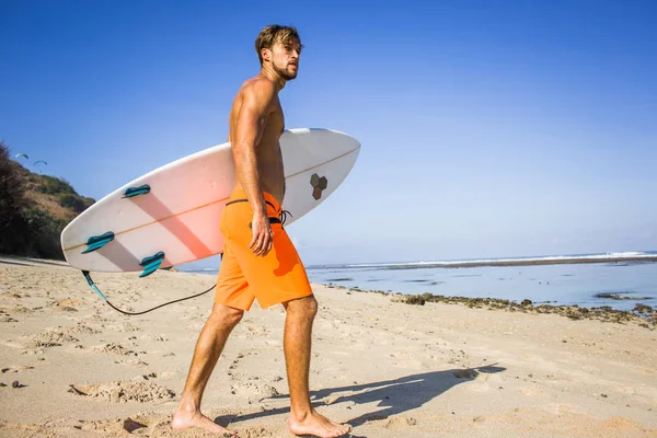 Side view of young athletic sportsman with surfing board walking on sandy beach — Stock Photo