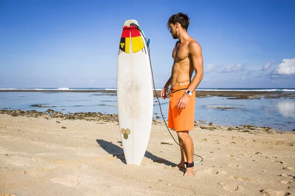 Young surfer looking at surfing board on sandy beach near ocean — Stock Photo