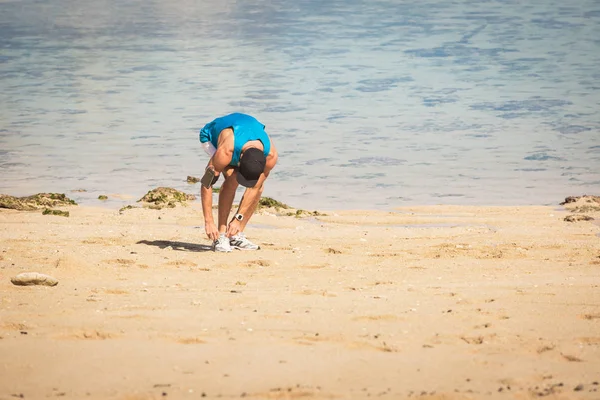 Deportista con brazalete de teléfono inteligente atando cordones en la playa cerca del mar - foto de stock