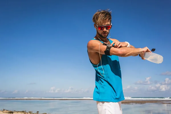 Atlético hombre con botella de deporte usando smartphone en brazalete cerca del mar - foto de stock