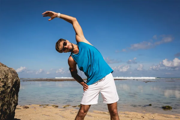 Sportsman in sunglasses stretching on beach near sea — Stock Photo