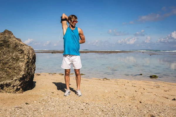 Handsome sportsman in sunglasses stretching arms on seashore — Stock Photo