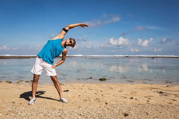 Homme athlétique en vêtements de sport s'étendant sur la plage près de la mer — Photo de stock