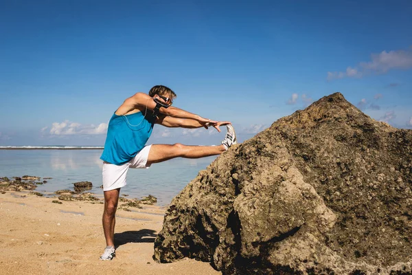 Sportif avec brassard étirant la jambe près du rocher sur la plage — Photo de stock