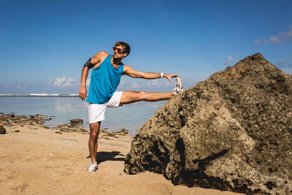 Sportive man stretching leg near rock on beach — Stock Photo