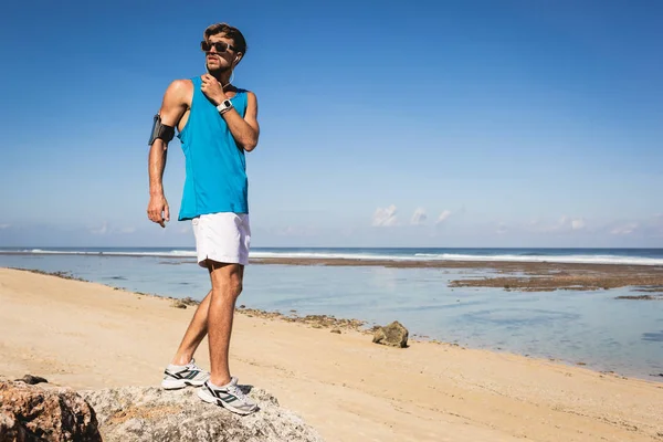 Sportsman listening music with smartphone in armband while standing on beach, Bali, Indonesia — Stock Photo