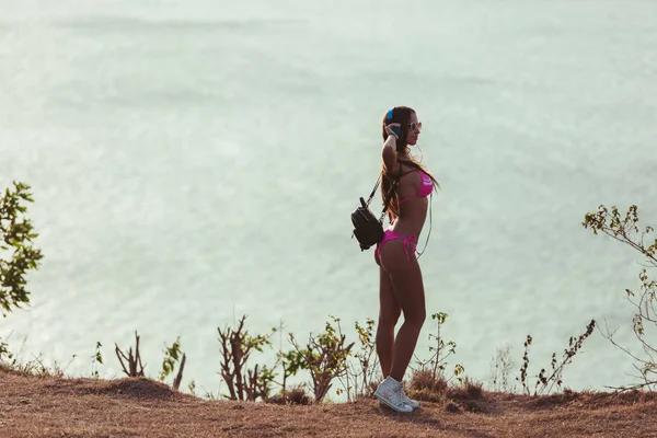 Vista lateral de chica atractiva en auriculares y bikini rosa de pie en la costa - foto de stock