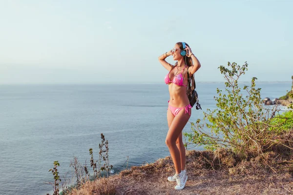 Jolie fille en bikini rose touchant écouteurs et regardant loin de la plage — Photo de stock