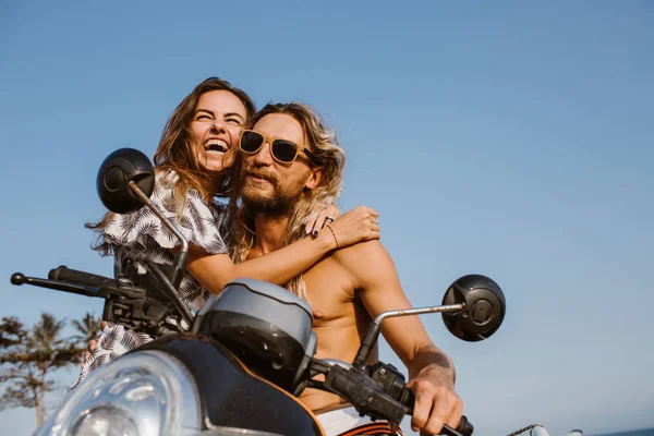 Vista de ángulo bajo de la pareja riendo abrazándose cerca de scooter en la playa en bali, indonesia - foto de stock