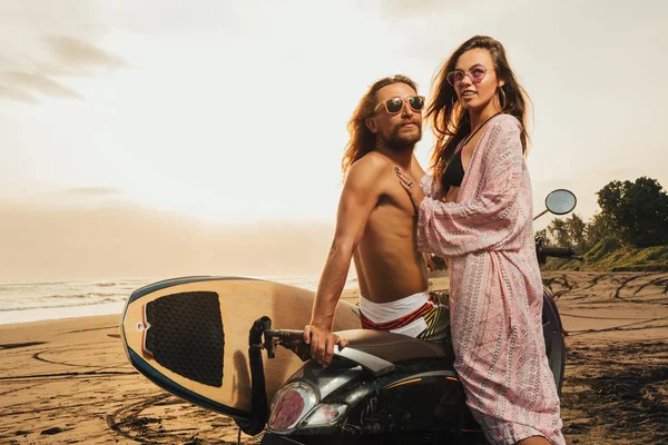 Sonriente pareja de pie cerca de scooter con tabla de surf en la playa durante el atardecer en bali, indonesia - foto de stock