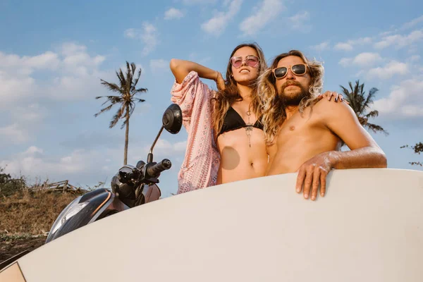 Baixo ângulo vista de casal de pé perto de scooter com prancha de surf na praia em bali, indonésia — Fotografia de Stock