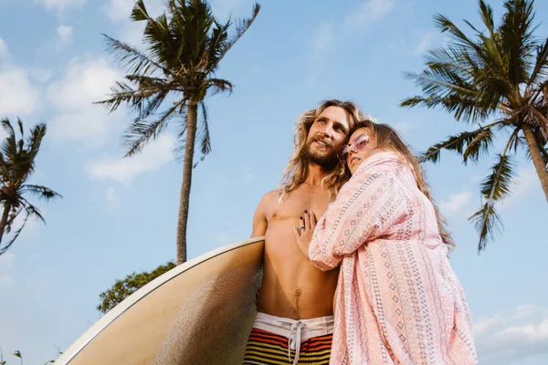 Vista de ángulo bajo de novia abrazando novio sonriente con tabla de surf en la playa en bali, indonesia - foto de stock