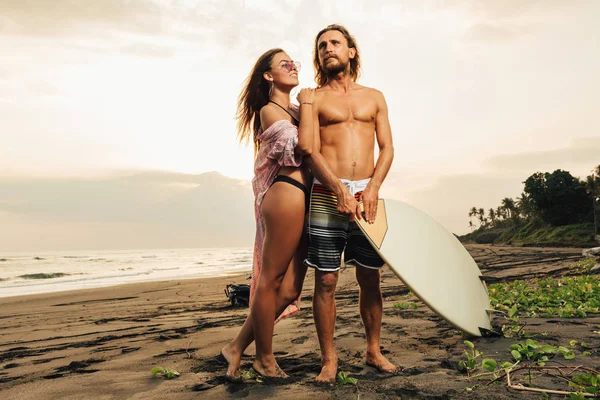 Fidanzata sorridente che abbraccia fidanzato con tavola da surf sulla spiaggia durante il tramonto a bali, indonesia — Foto stock