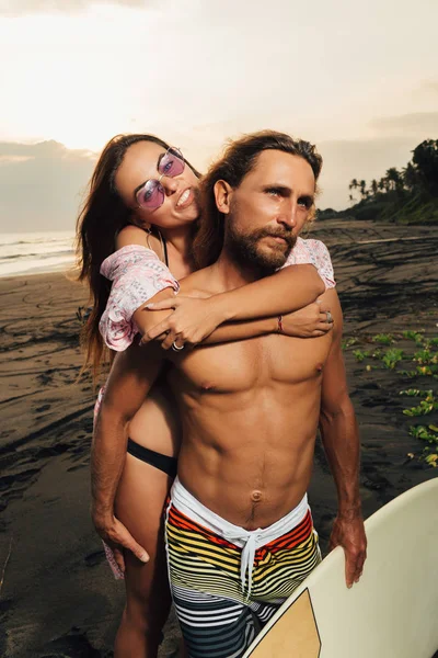 Petite amie souriante étreignant petit ami avec planche de surf sur la plage à Bali, indonesia — Photo de stock