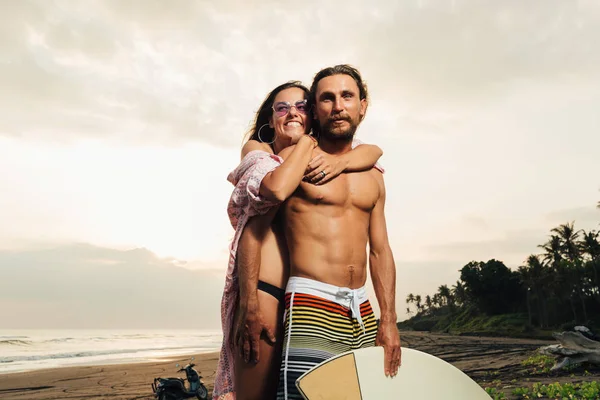 Fidanzata sorridente che abbraccia fidanzato con tavola da surf sulla spiaggia di bali, indonesia — Foto stock