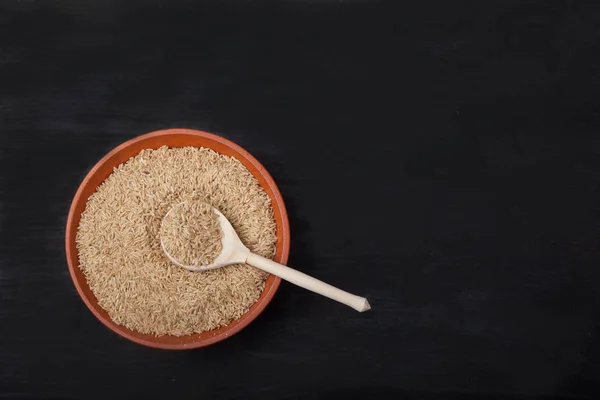 Unpolished rice with a wooden spoon in a clay bowl on a black ba — Stock Photo, Image