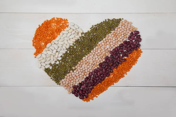 Fagioli, lenticchie e fagioli disposti a forma di cuore su uno sfondo di legno bianco Foto Stock